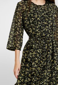 Robe noire à motifs floraux jaunes et blancs, en tissu semi-transparent, à manches trois quarts et avec une taille ceinturée.