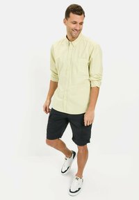 Jasnożółta koszula z długim rękawem i kieszonką w stylu button-up, zestawiona z czarnymi szortami cargo i białymi trampkami z czarnymi akcentami. Swobodny krój.