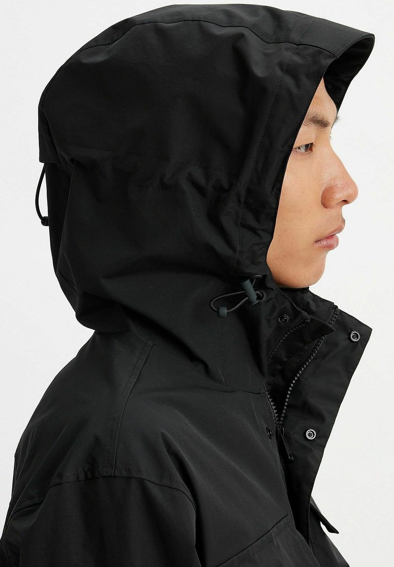 Veste à capuche noire avec une finition en nylon. Comprend des cordons de serrage ajustables, des fermetures à pression et une poche avant zippée. Profil latéral affiché.