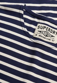 Encolure de chemise rayée horizontale bleu marine et blanc avec étiquette blanche cousue indiquant "Superdry Crafted Merchant Store M Made in India."