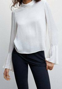 Blouse blanche à manches longues avec un col haut et une texture plissée. Présente des poignets évasés et une coupe décontractée, portée avec un pantalon foncé.