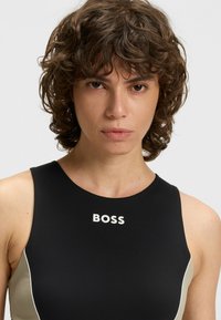 Schwarze Badebekleidung mit Rundhalsausschnitt, versehen mit einem hellbraunen Seitenpanel und einem weißen "BOSS"-Logo auf der Brust. Glattes, eng anliegendes Material.