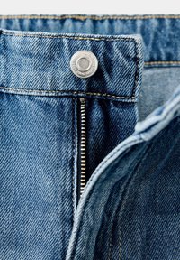 Gros plan sur la fermeture éclair d'un jean en denim bleu partiellement ouverte avec un bouton argenté fixé à la ceinture.