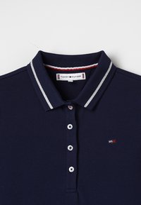 Granatowa koszulka polo z teksturowanej tkaniny, z prążkowanym kołnierzykiem z srebrnymi paskami, trzema guzikami i małym logo na klatce piersiowej.