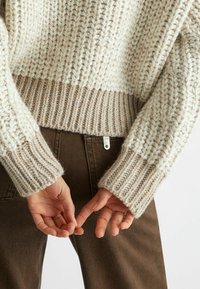 Maglione a maglia grossa in crema e beige con polsini a coste. Presenta un filato testurizzato e un'etichetta visibile all'orlo. Mani posizionate dietro.