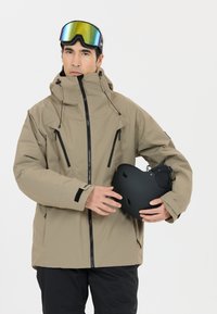 Beige skijas met een hoge kraag, zwarte accenten en ventilatiedetails, vastgehouden met een zwarte skiketting. Zwarte broeken maken de outfit compleet.