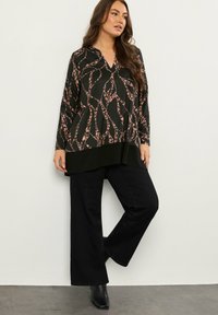 Schwarzes Langarmoberteil mit V-Ausschnitt, das mit einem pinken und beigen abstrakten Muster verziert ist, kombiniert mit schwarzen weit geschnittenen Hosen und Absatzstiefeln.