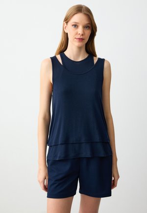 Jonge vrouw in een marineblauwe gelaagde mouwloze top en bijpassende shorts die tegen een plain witte achtergrond staat.