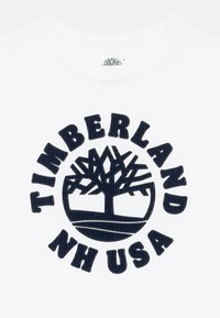 Hvit bomull t-skjorte med en stor marineblå sirkulær logo som har teksten "TIMBERLAND NH USA" og et tre-design i midten.
