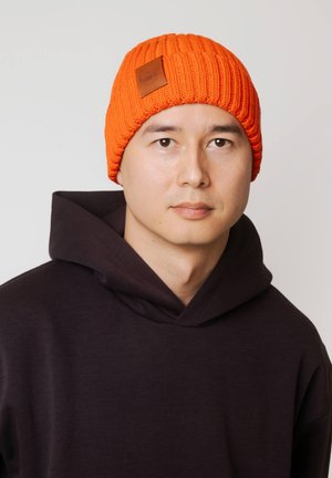 Pomarańczowa dziergana czapka z prążkowaną fakturą i brązową skórzaną naszywką. Noszona z ciemną bluzą z kapturem, na tle jednolitej białej ściany.