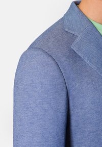 Světle modrý blejzr s texturovanou tkaninou, ostrými klopami a strukturovanými rameny. Detailní záběr zdůrazňuje vzor tkaní.
