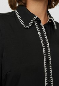 Black shirt with a white embroidered collar and button placket, featuring bold stitching and smooth texture.