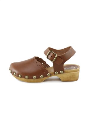 Zueco de cuero marrón con borde festoneado, tachuelas de latón, tacón de madera y correa ajustable en el tobillo con hebilla de latón sobre fondo blanco.