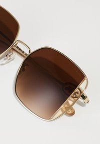 Lunettes de soleil carrées teintées marron avec une monture en métal doré, dotées d'un coussinet de nez transparent et de bords sculptés pour un design moderne.