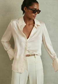 Blusa de seda crema con cuello clásico, mangas largas y puños con botones, combinada con pantalones de talle alto beige claros y un cinturón delgado.