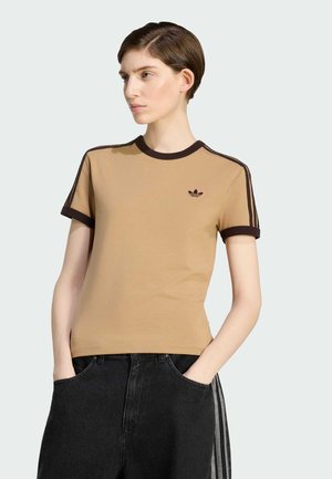 Kobieta o krótkich brązowych włosach, ubrana w beżowy t-shirt Adidas z ciemnym wykończeniem oraz czarne spodnie z paskami po bokach, ręce w kieszeniach, patrzy w lewo.