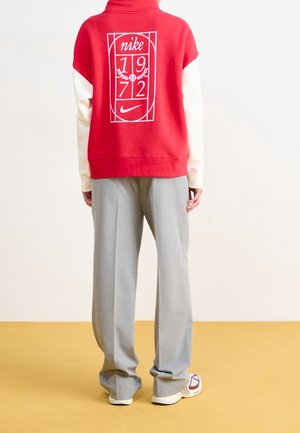 Person iført en rød og hvid Nike sweatshirt med "1972" logo på ryggen, grå vidde bukser og hvide sneakers, der står på gul gulv.