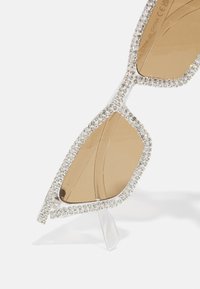Selged prillid geomeetrilise kujuga, mille raam on kaunistatud rhinestones'iga ja pruunide toonitud läätsedega. Tekstuuriga selged käed.