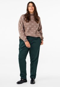 Hnedý textúrovaný sveter s bielymi vzormi, skombinovaný so zelenými zúženými nohavicami. Čierne mokasíny dotvárajú celý outfit.