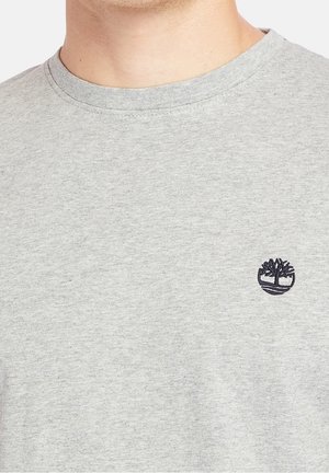 Zbliżenie osoby noszącej jasnoszary t-shirt z okrągłym dekoltem i małym czarnym logo drzewa na lewej piersi.