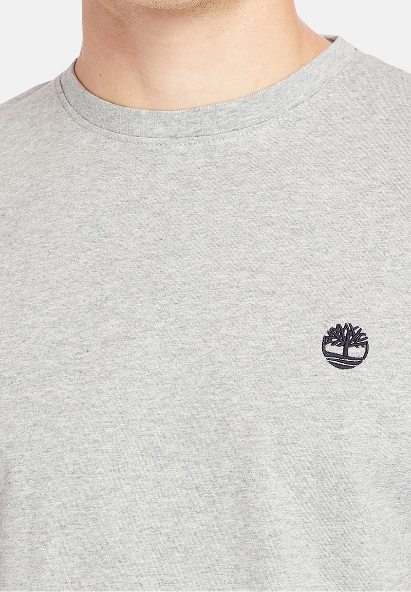Gros plan d'une personne portant un t-shirt gris clair à col rond avec un petit logo arbre noir sur la poitrine gauche.