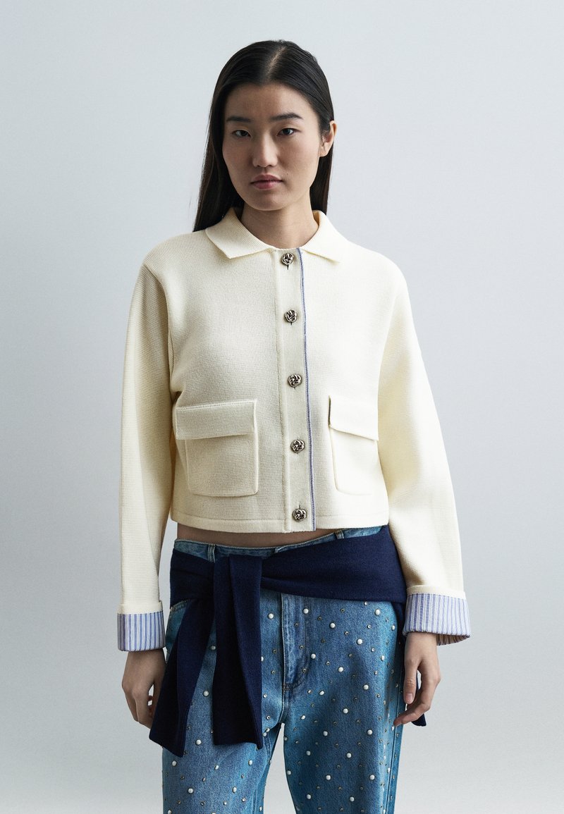 Jeune femme portant une veste crème boutonnée avec des poches avant, des poignets rayés retroussés et un jean bleu à clous, avec un pull marin noué autour de la taille.