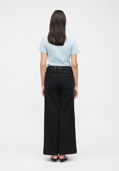 Γυναίκα με μακριά καστανά μαλλιά στέκεται με την πλάτη, φοράει ένα ανοιχτό μπλε κοντομάνικο πουκάμισο, μαύρο παντελόνι με ψηλή μέση και ευρύ ποδόγυρο, και μαύρα σανδάλια.