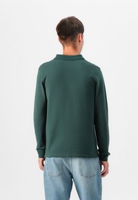 Tummanvihreä pitkähihainen t-paita, jossa on kaulus, sileä pinta ja rento istuvuus, yhdistettynä vaaleansinisiin farkkuihin.