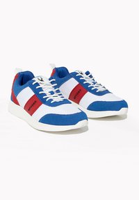 Blå och vita sportskor med röda accenter. De har en ovandel i mesh, snören och en texturerad gummisula. Varumärkeslogotyp på sidan.