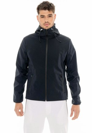 Homme portant une veste noire à capuche, zippée, avec des cordons et des poches, assortie à un pantalon blanc, sur un fond blanc uni.