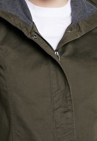 Chaqueta verde oliva con un cuello de forro polar gris, que cuenta con un cierre frontal y detalles cosidos en una tela de mezcla de algodón.
