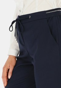 Kobieca dłoń spoczywa na granatowych spodniach z elastycznym pasem, w połączeniu z białą, długą koszulą z mankietami i guzikiem z kryształkami.