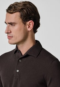 Dunkelbraunes Poloshirt aus Baumwolle mit klassischem Kragen, Zwei-Knopf-Leiste und feiner Struktur. Verstellbare Knöpfe setzen zusätzliche Akzente.