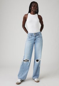 Jean délavé bleu clair avec une coupe large et des déchirures au niveau des genoux, associé à un débardeur blanc à côtes et des baskets décontractées.