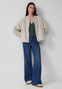 Femme portant une veste zippée beige, un top vert foncé en V, un jean bleu large et des baskets blanches, se tenant devant un mur uni.