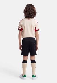 Child with curly hair stands facing away, wearing beige and red sports shirt, black shorts, beige knee-high socks, and white sneakers with green heels.