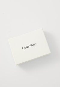 Boîte rectangulaire blanche inclinée en diagonale avec le logo noir "Calvin Klein" centré sur le couvercle, sur fond blanc uni.