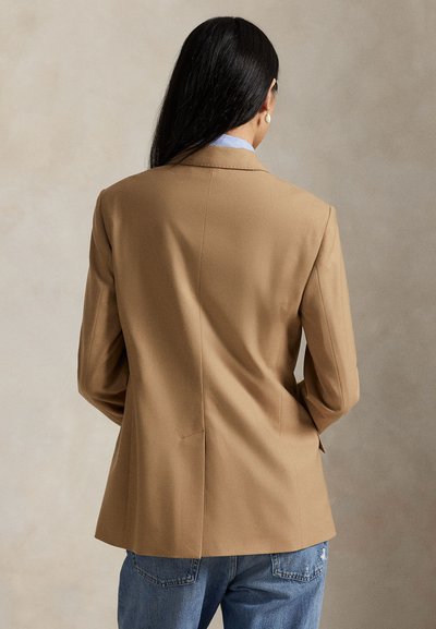 Béžový blazer so štruktúrovaným dizajnom, s jedným rozventilovaním a prispôsobeným strihom. Nosený cez svetlomodrú košeľu a kombinovaný s džínsami.