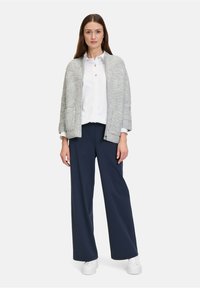 Cardigan aperto grigio chiaro a maglia larga con due tasche anteriori, indossato sopra una camicia bianca con bottoni e pantaloni larghi blu navy, abbinato a sneakers bianche.