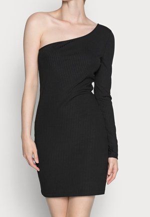 Femme portant une robe noire moulante côtelée avec une manche longue et une épaule nue, debout devant un fond clair uni.