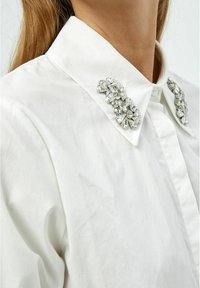Camicia bianca con colletto decorato da strass trasparenti e scintillanti. Tessuto liscio, design del colletto pieghevole e finitura nitida.