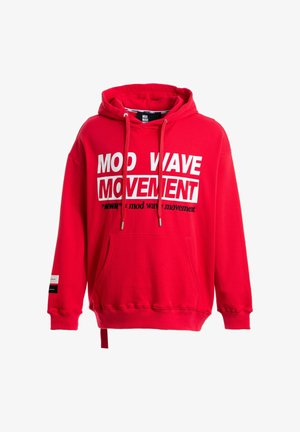 Joven de pie con una sudadera roja con capucha con el texto "MOD WAVE MOVEMENT", pantalones blancos con una correa colgante y botas marrones sobre un fondo blanco.