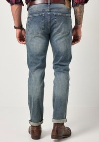 Jeans de mezclilla azul claro, corte recto, ligeramente desgastados, con puños doblados y detalles en el bolsillo trasero. Llevados con botas marrones.