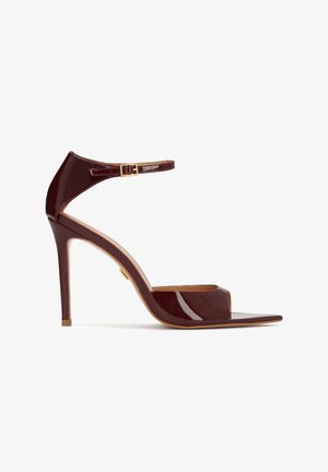 Tmavá bordová sandála na vysokom podpätku z lesklého laku, s remienkom okolo členka a zlatou prackou, otvorenou špičkou a úzkym ihličkovým podpätkom.