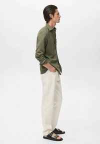 Camisa verde oliva de botones, pantalones crema y sandalias negras con dos hebillas, mostradas de lado contra un fondo liso.