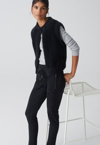 Gilet sans manches noir en tissu pelucheux avec un col rond superposé sur un haut à manches longues gris clair, associé à un pantalon noir ajusté.