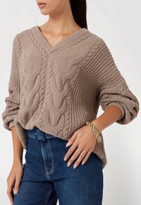 Beige gebreide trui met een V-hals, met textuurrijke kabelpatronen, losse mouwen, gedragen over donkerblauwe jeans. Gouden armband om de pols.