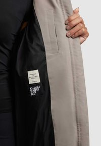 Light gray jacket with a black inner lining, featuring two labels on the left side and a pocket on the inside. Smooth fabric texture.