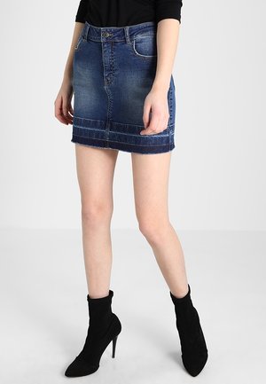 Femme portant une mini-jupe en jean bleu avec ourlet effiloché, un top noir à manches longues et des bottines noires à talons, debout sur un fond uni.