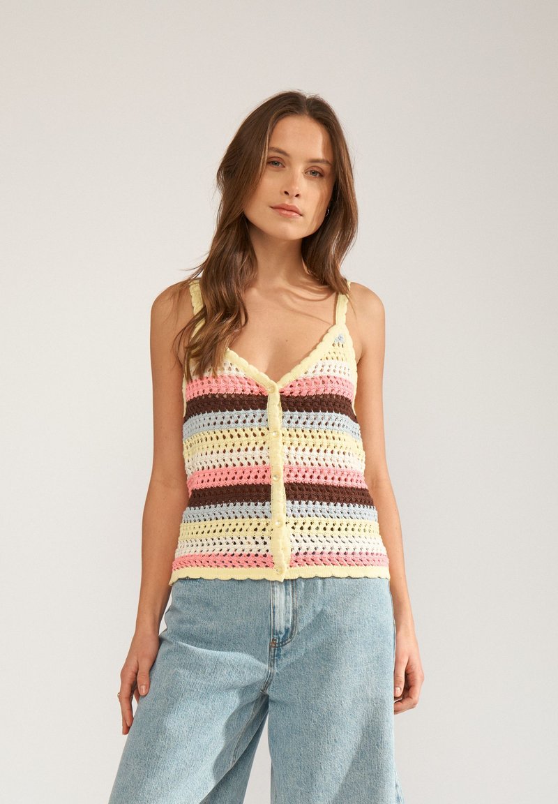 Femme portant un débardeur crocheté à rayures multicolores avec des boutons, associé à un jean taille haute bleu clair, debout devant un fond uni.